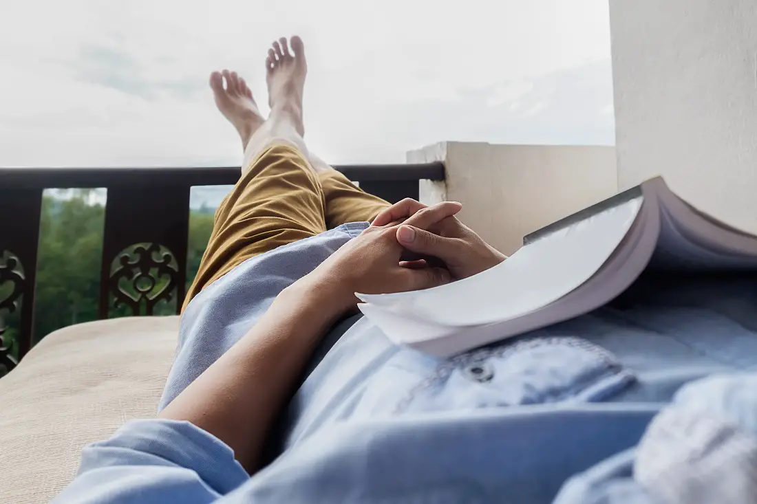 Young man reading a book lying on soft mattress in relaxing bed at terrace with green nature view. Fresh air in the morning of weekend or free day. Relax or education background idea. Selective focus.